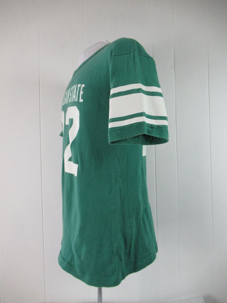 "Vintage 1970s or 80s college t shirt. Made of green cotton with thick white college graphics: Michigan State #22. Has Champion blue block label. Size large. Actual measurements are: 39\" around the chest 39\" around the waist 20\" shoulder seam to shoulder seam 28\" overall length In very good condition with 3\" of seam repair to the cuff." Retro College T-shirt With Screen Print, Retro Crew Neck T-shirt For School, Green Short Sleeve T-shirt For College, Retro Green T-shirt For Fans, Retro Green T-shirt For Streetwear, Collegiate Green T-shirt With Logo Print, Vintage College T-shirt With Graphic Print, Varsity Cotton Tops For School, Cotton Varsity Tops For School