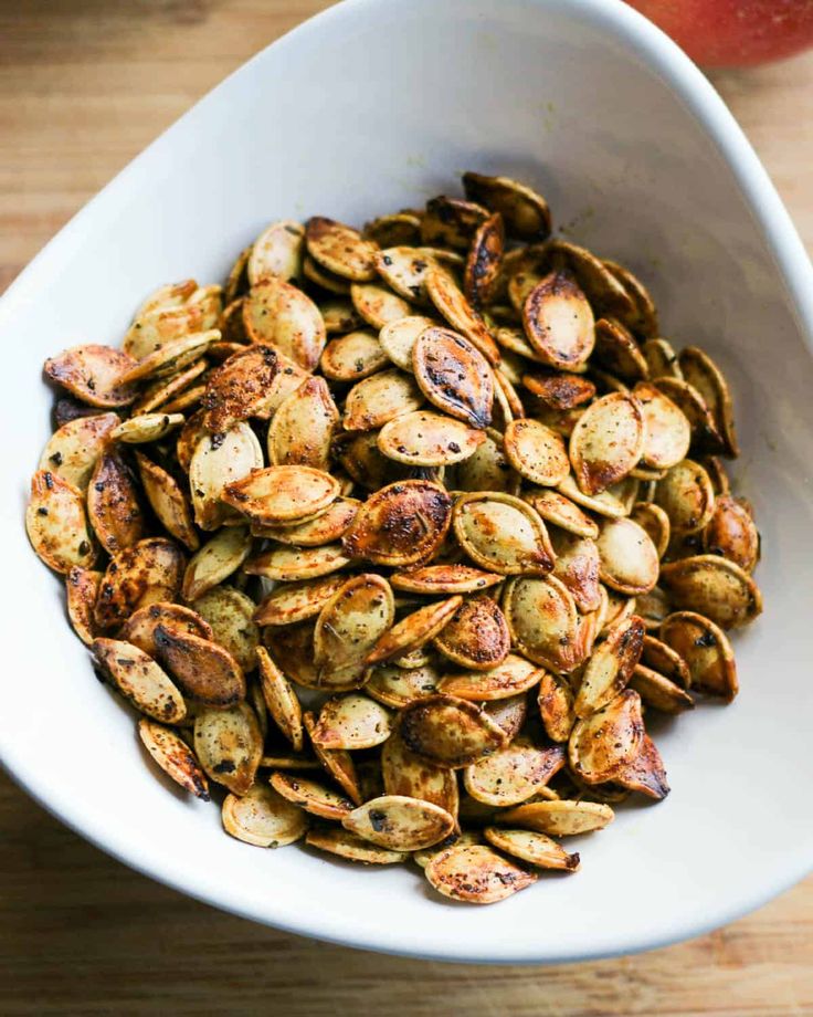 roasted pumpkin seeds in a white bowl on a wooden table with an apple behind it