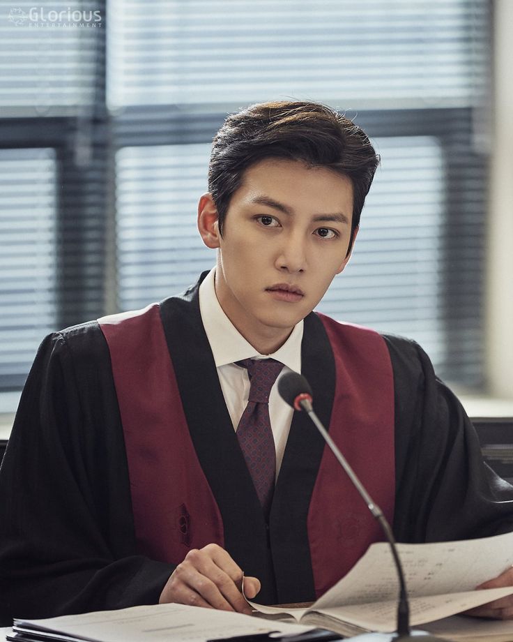 a young man in a graduation gown sitting at a desk with papers and a microphone