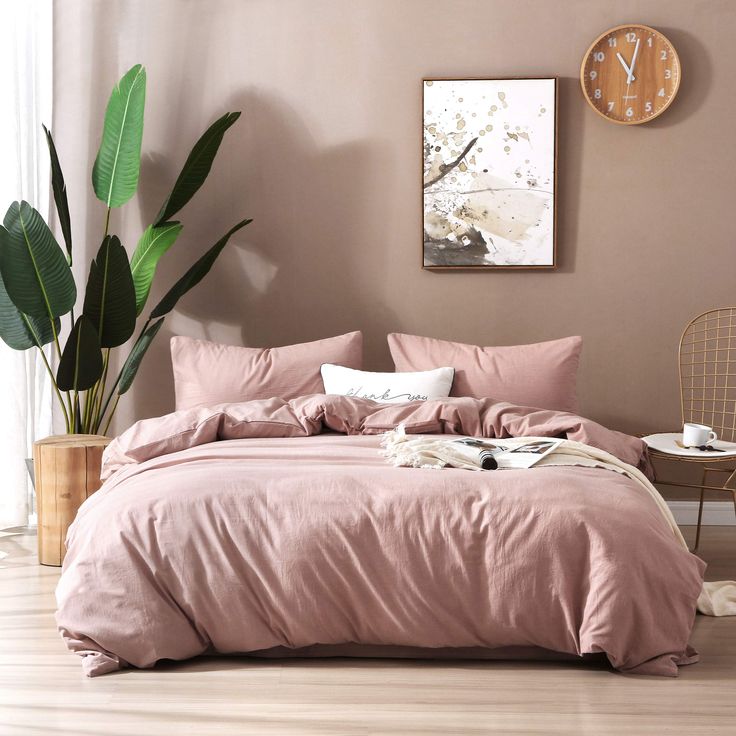 a bed with pink sheets and pillows next to a potted plant in front of a window