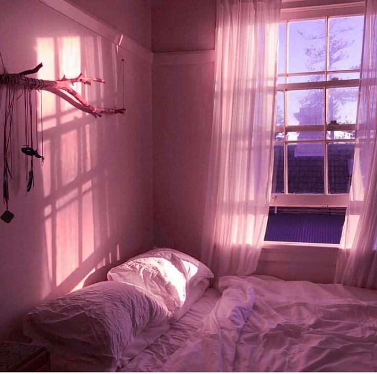 an unmade bed in front of a window with pink light coming through the curtains