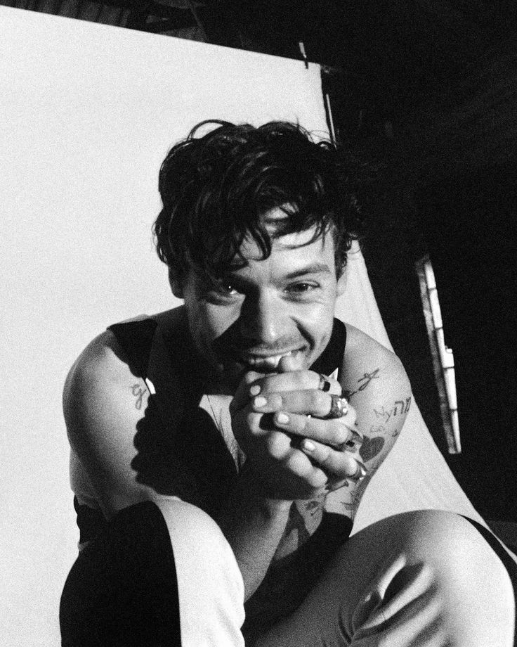 a black and white photo of a man with tattoos on his arm, sitting in front of a wall