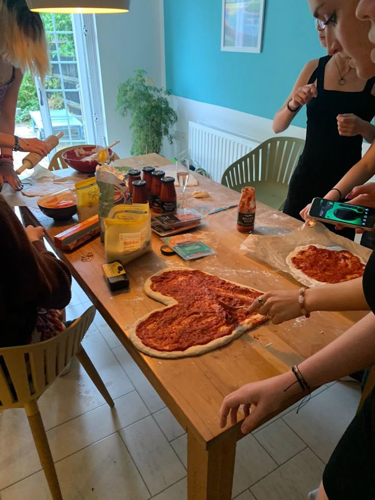 three women are preparing pizzas on a table in a kitchen with blue walls and white tile flooring