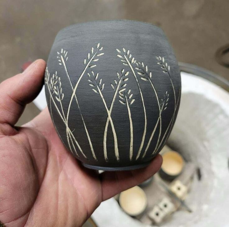 a hand holding a black and white vase with plants painted on it's side