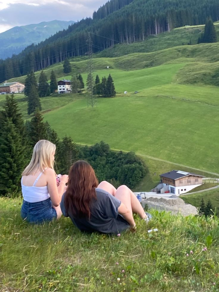 two women sitting on top of a hill looking at the valley and houses in the distance