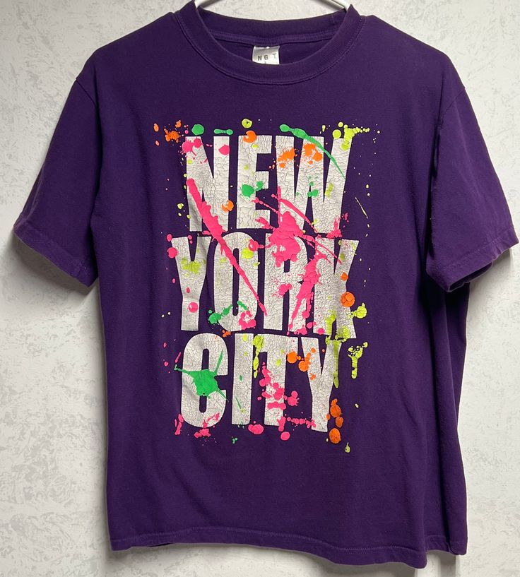 Purple tee shirt features short sleeves and New York City graphic with neon splatter accents. This item is old and it shows some signs of wear. The graphic is crackled and there is some pilling and discoloration in the underarm area (see photos). This item is listed as an adult M and is in good vintage condition with no holes. Purple Grunge T-shirt With Letter Print, Multicolor Letter Print Grunge T-shirt, Multicolor Grunge T-shirt With Letter Print, Urban Purple Short Sleeve T-shirt, Casual Paint Splatter T-shirt For Streetwear, Casual T-shirt With Paint Splatter For Streetwear, Urban Style Purple Short Sleeve T-shirt, Short Sleeve Graphic Tee With Paint Splatter, Spring Crew Neck T-shirt With Paint Splatter