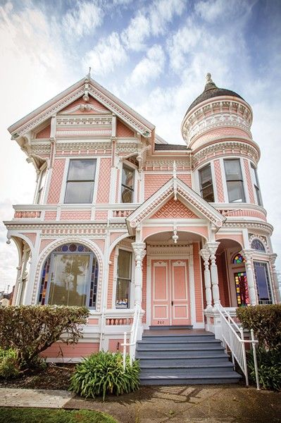 a pink and white victorian style house with stairs leading up to the front door on a sunny day