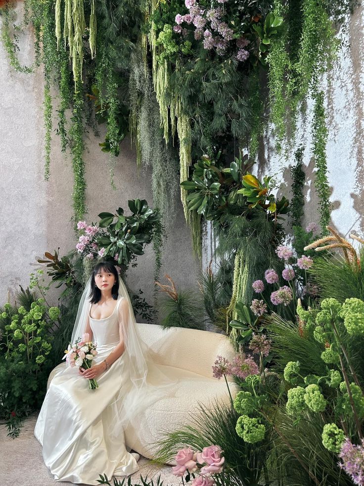 a woman in a wedding dress sitting on a couch surrounded by flowers and greenery