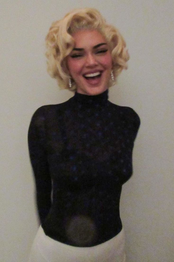 a woman with blonde hair wearing a black top and white skirt smiling at the camera