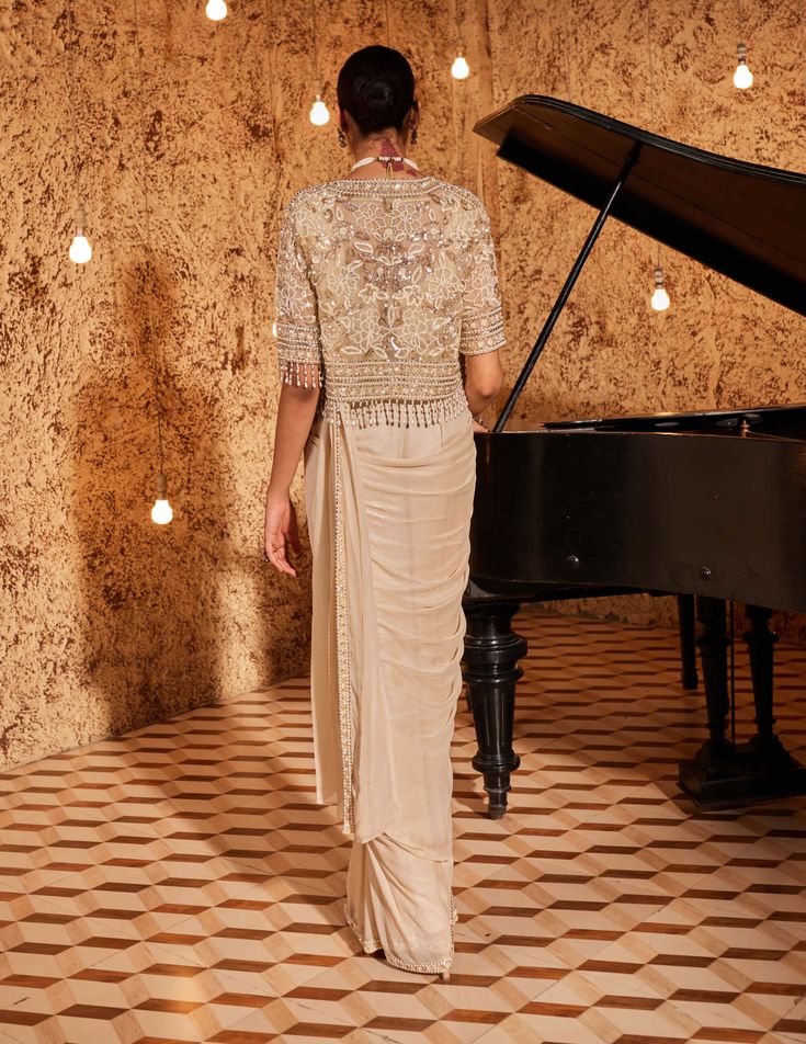 Embrace opulence in this golden ensemble featuring a short cape jacket adorned with intricate floral embroidery. The drape sari is elegantly finished with an embroidered border and a coordinating belt, adding a touch of glamour. The padded blouse, with a concealed zip on the back, ensures a seamless fit. Step into luxury with this gorgeous outfit that seamlessly blends traditional elegance with contemporary style. Festive Draped Sharara With Sheer Dupatta, Reception Choli With Cutdana And Cape Sleeves, Festive Draped Lehenga With Resham Embroidery, Sharara With Cutdana And Cape Sleeves For Reception, Sharara With Cutdana For Reception, Festive Draped Resham Embroidery Lehenga, Festive Sharara With Cutdana And Cape Sleeves, Diwali Sharara With Cutdana And Cape Sleeves, Navratri Dupatta With Cutdana And Cape Sleeves