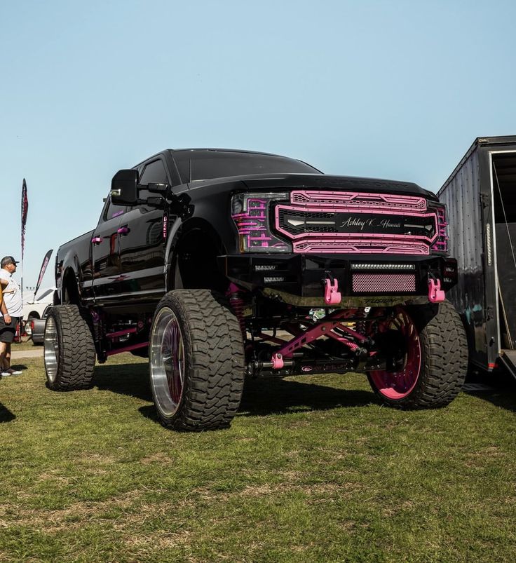 a large black truck with pink wheels parked in the grass next to other trucks and people