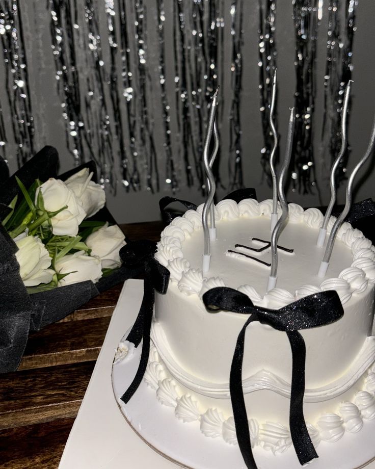 a white cake with black ribbon and forks on it sitting next to some flowers in front of silver sequins