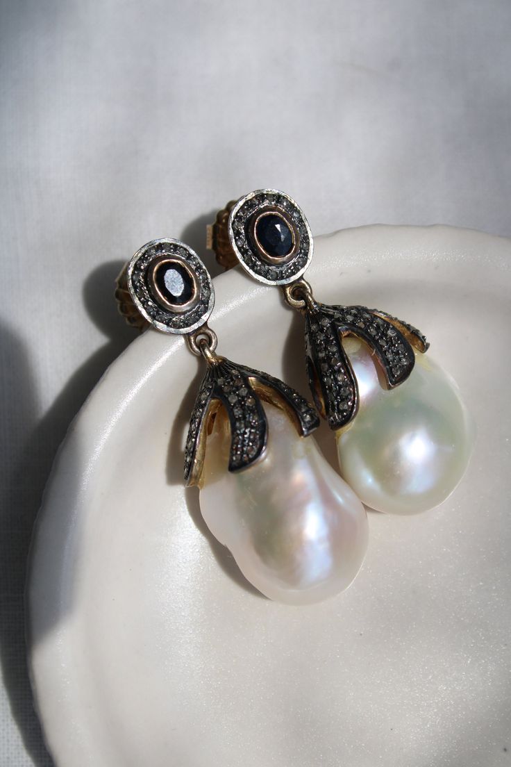 This is an incredible one-of-a-kind find with these Victorian pearl earrings, a true testament to timeless elegance and sophisticated craftsmanship. Each earring boasts a lustrous baroque pearl nestled within a setting of silver-topped gold, adorned with high domed rose cut diamonds that catch the light for a subtle, enchanting shimmer. The center stones, one a deep black onyx and the other a rich blue sapphire, hint at their possible origins as a bridal accessory, adding a touch of romantic his Luxury Oval Pearl Earrings, Luxury Pear-shaped Earrings For Evening, Luxury Oval Jewelry For Opera, Luxury Teardrop Earrings With High Luster, Luxury Teardrop Pearl Earrings With Diamond Accents, Luxury Evening Drop Pearl Earrings, Luxury Drop Pearl Earrings For Evening, Luxury Rose Cut Pear Shaped Jewelry, Timeless Rose Cut Diamond Earrings