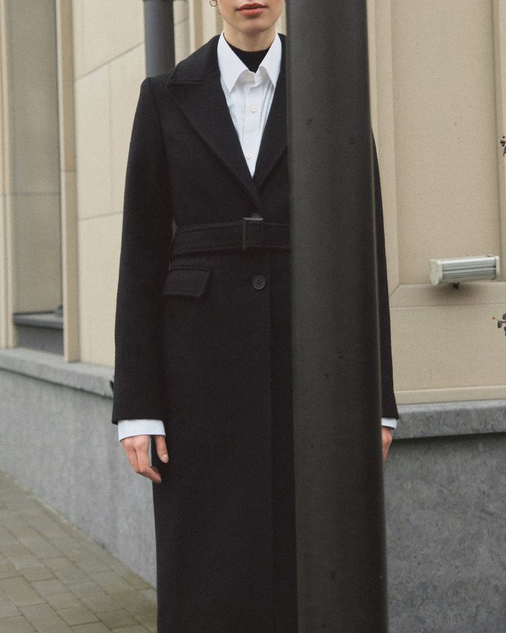 a woman standing next to a pole in front of a building wearing a black coat and white shirt