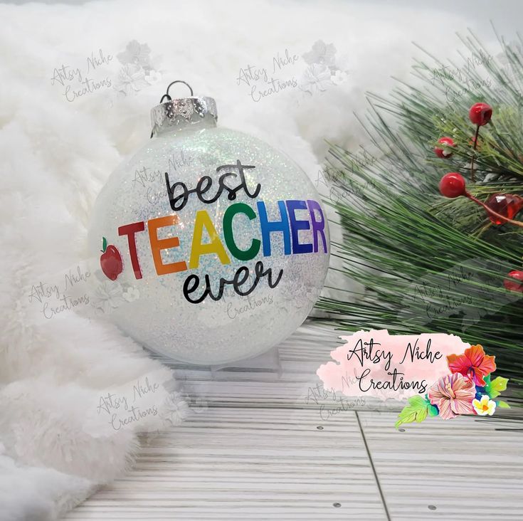 a white ornament with the words best teacher ever on it sitting next to a pine tree