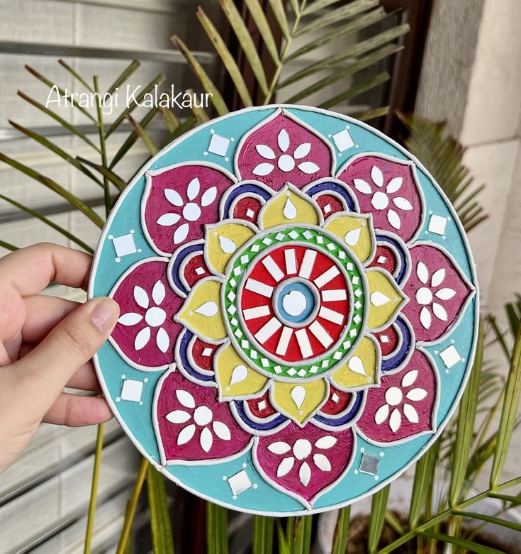 a hand holding up a colorful plate with designs on the front and side, in front of a plant