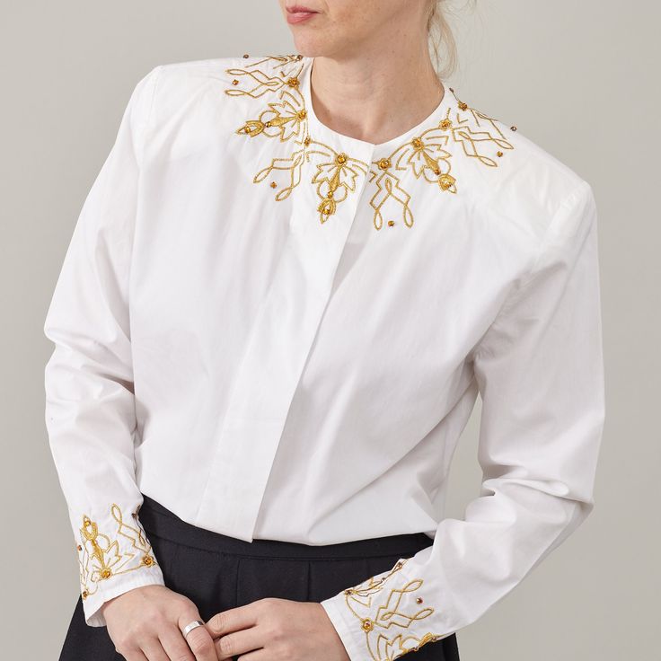 "Indulge in timeless glamour with our White Cotton Shirt adorned with Golden Embroidery Details. This elegant festive blouse is a statement piece crafted to elevate your ensemble effortlessly. Designed with meticulous attention to detail, it features hidden button placket and subtly padded shoulders, offering a refined silhouette that exudes sophistication. KEY FEATURES: - Luxurious white cotton fabric - Intricate golden embroidery details for a touch of opulence - Hidden buttons placket for a sleek finish - Subtly padded shoulders for added structure and elegance - Suitable for women size S - M - Excellent vintage condition - Dry clean only Fabric Information: 100% cotton MEASUREMENTS (while lying flat): Armpit to armpit: 52 cm | 20.5\" Length: 67 cm | 26.4\" Sleeves: 57 cm | 22.4\" Shoul Embroidered Long Sleeve Formal Tops, Embroidered Long Sleeve Tops For Formal Occasions, Embellished Fitted Shirt For Workwear, Elegant Long Sleeve Shirt With Embellished Collar, White Embellished Top For Work, Luxury Tops With Embellished Collar, Spring Formal Tops With Embellished Collar, Elegant Long Sleeve Embroidered Shirt, Spring Formal Top With Embellished Collar