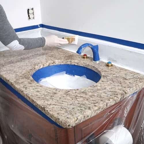 a woman is painting a bathroom sink with blue tape on the counter top and around it