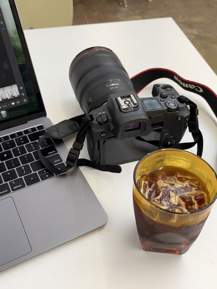 an open laptop computer sitting on top of a white table next to a drink and camera