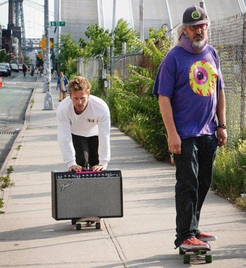 a man riding a skateboard down a sidewalk next to another man with a suitcase