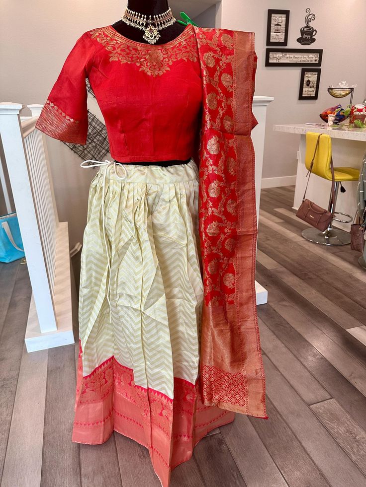 This is a beautiful, fully stitched mixed banarasi silk half saree set for girls/young women. The lehenga is bordered by a detailed jacquard border and is a golden beige shade with chevron zari design on the body.  Lehenga comes with drawstring and a zipper at the closure and is lined. The set comes with a back open, padded blouse with doris at the back and is a red shade. Stitched blouse size is 36 inches bust and has room inside for adjustment unto size 38 inches. The blouse can also be altered to a smaller size upon request. Length of lehenga - 39.5 inches Length of blouse sleeves - 11 inches Bust size of blouse - 36 inches and adjustable to smaller/bigger (unto 38 inches) size Size S | Fully stitched Banarasi mixed silk half saree set | girls half saree set online shopping usa | leheng Silk Anarkali Set With Tilla, Silk Anarkali Set With Tilla Detailing, Unstitched Tilla Sets For Diwali, Silk Sets With Tilla Details For Diwali, Silk Tilla Sets For Diwali, Navratri Tussar Silk Sets With Cutdana, Chanderi Sets With Tilla For Navratri, Multicolor Banarasi Silk Dupatta With Tilla, Navratri Chanderi Sets With Tilla Detailing