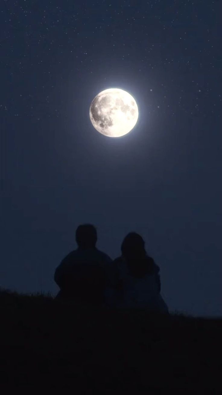 two people sitting on a hill watching the moon