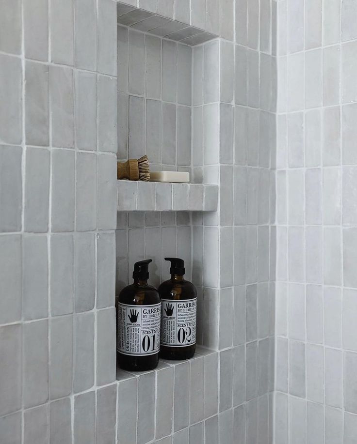 two bottles are sitting in the corner of a white tiled shower stall with shelves above them