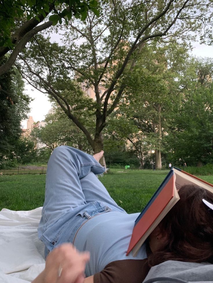 a person laying down reading a book in a park with trees and grass behind them