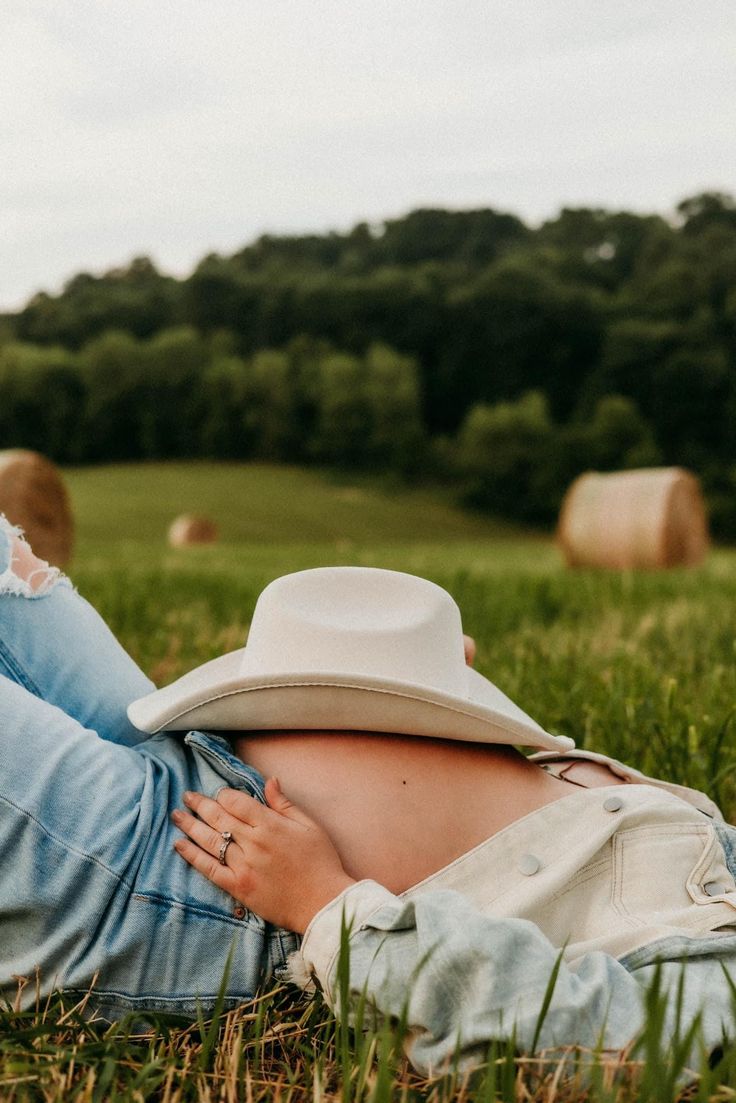 a woman laying in the grass wearing a white hat and blue jeans with her hands on her stomach