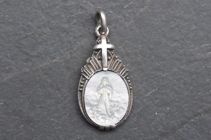 Art Deco pendant crafted from 835 silver, featuring a meticulously carved mother of pearl depiction of the Virgin Mary.  The pendant is adorned with elegant milgrain detailing and topped with a cross, the reverse is marked "835," attesting to its silver purity.  In very good condition, this piece is a stunning representation of both spiritual reverence and vintage craftsmanship, making it a perfect addition to any jewelry collection or a thoughtful gift for someone special. Measures 1.2cm x 2.1cm - whitout bail, weights 1.6 grams. Let me know if you would like further photos or information. Thanks for your visit! Engraved Silver Mother Of Pearl Necklace, Elegant White Miraculous Medal Jewelry, Victorian Engraved White Jewelry, White Engraved Victorian Jewelry, Antique Engraved White Jewelry, Antique White Jewelry With Polished Finish, Antique White Engraved Jewelry, Victorian Style White Engraved Jewelry, Silver Medallion With Mother Of Pearl