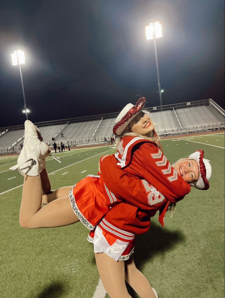 two cheerleaders in red and white outfits on a football field at night with stadium lights behind them