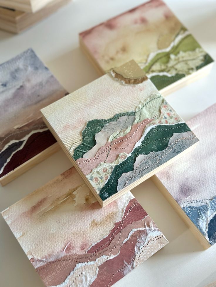 four different colored paintings sitting on top of a white table next to wooden blocks with lace