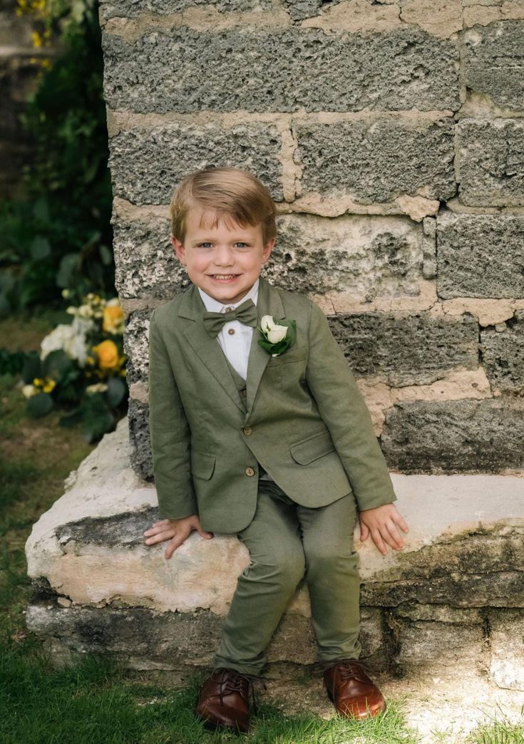 a young boy in a green suit and bow tie sitting on a stone bench with flowers