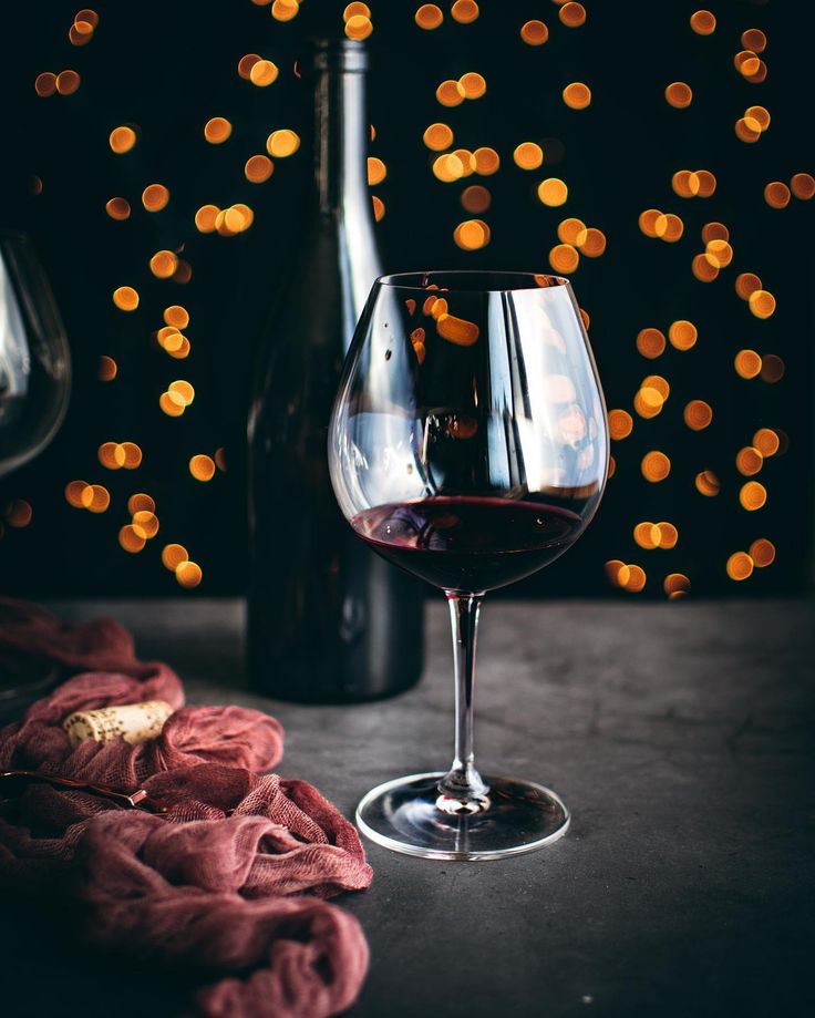 a glass of red wine next to a bottle of wine on a table with lights in the background