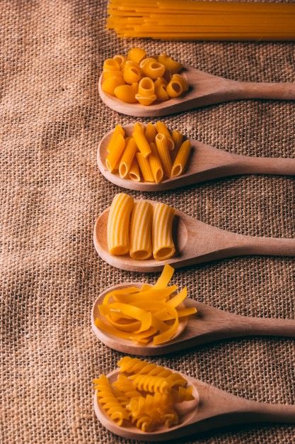 four wooden spoons filled with different types of pasta on top of a cloth covered table