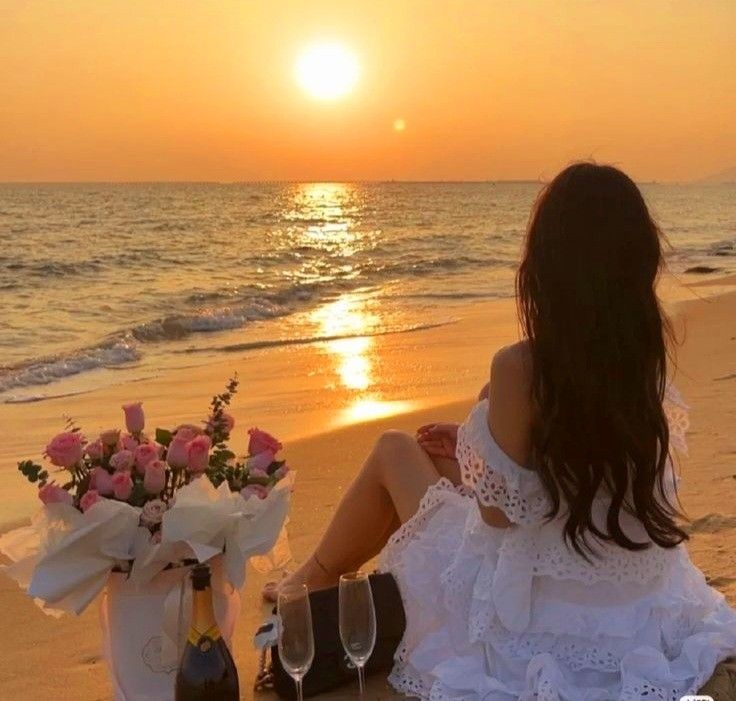 a woman sitting on the beach with two wine glasses in front of her and a vase full of flowers