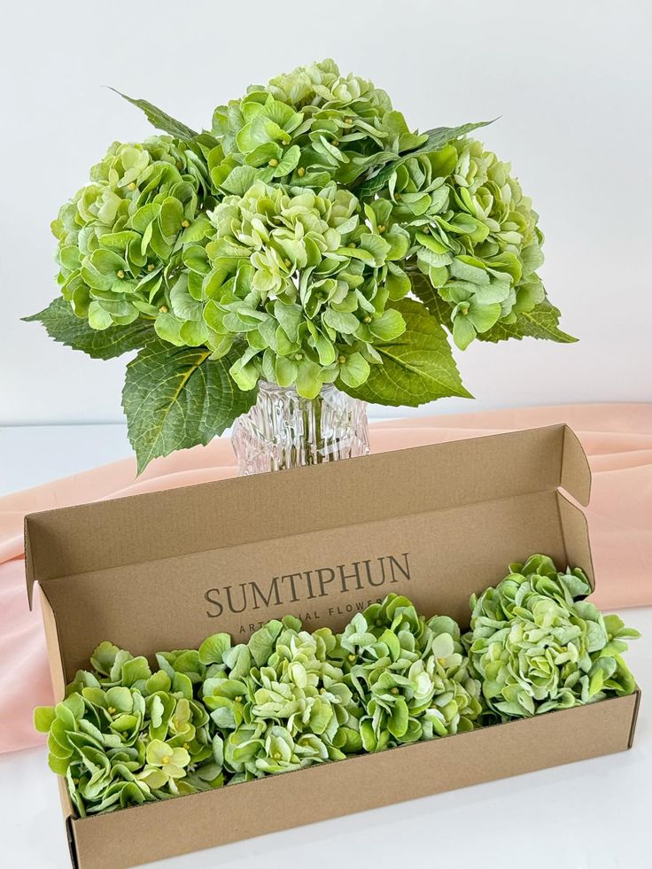 a box filled with green flowers sitting on top of a white table next to a pink cloth