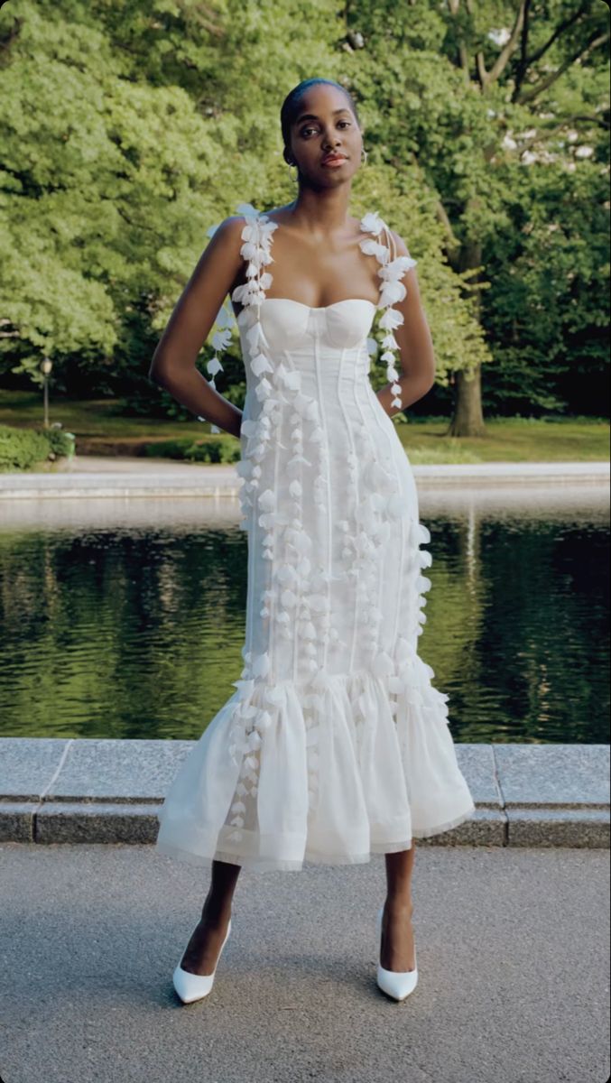 a woman standing in front of a pond wearing a white dress with ruffles