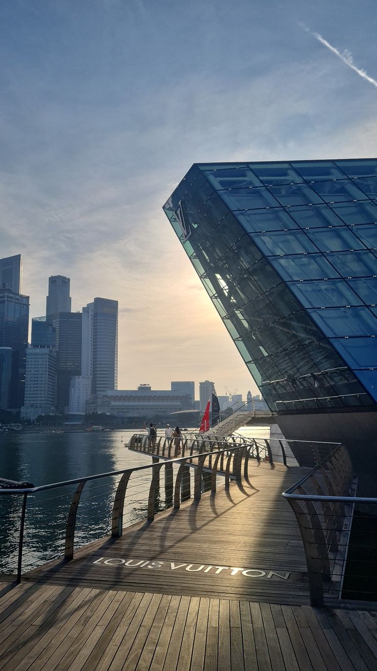 people are walking on the boardwalk next to the water in front of some tall buildings