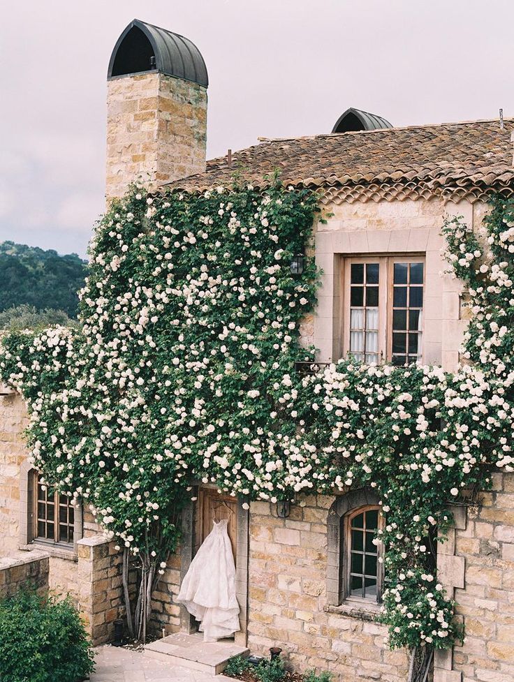 an old building with white flowers growing on it's side and a wedding dress hanging out the window