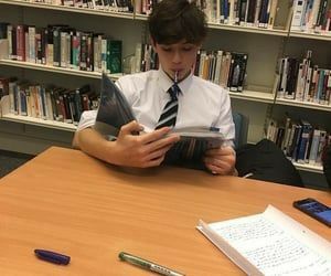 a young man sitting at a table in front of a bookshelf reading a book