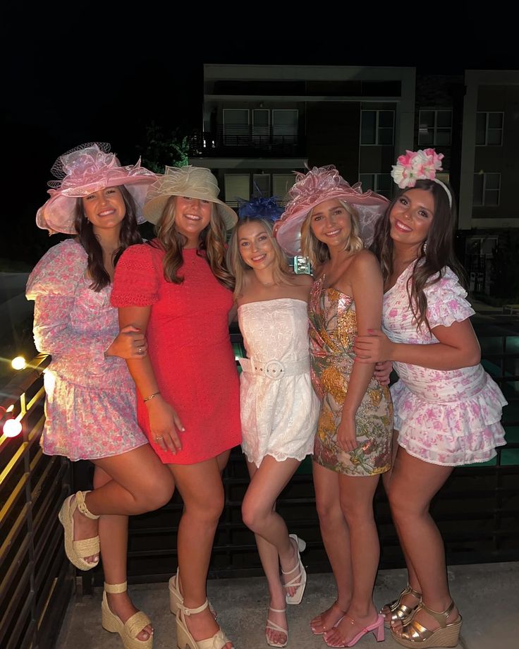 four women in dresses and hats posing for the camera at an outdoor event with lights behind them