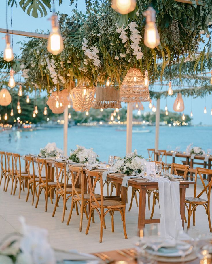 tables and chairs are set up for an outdoor wedding reception with hanging lights, flowers and greenery