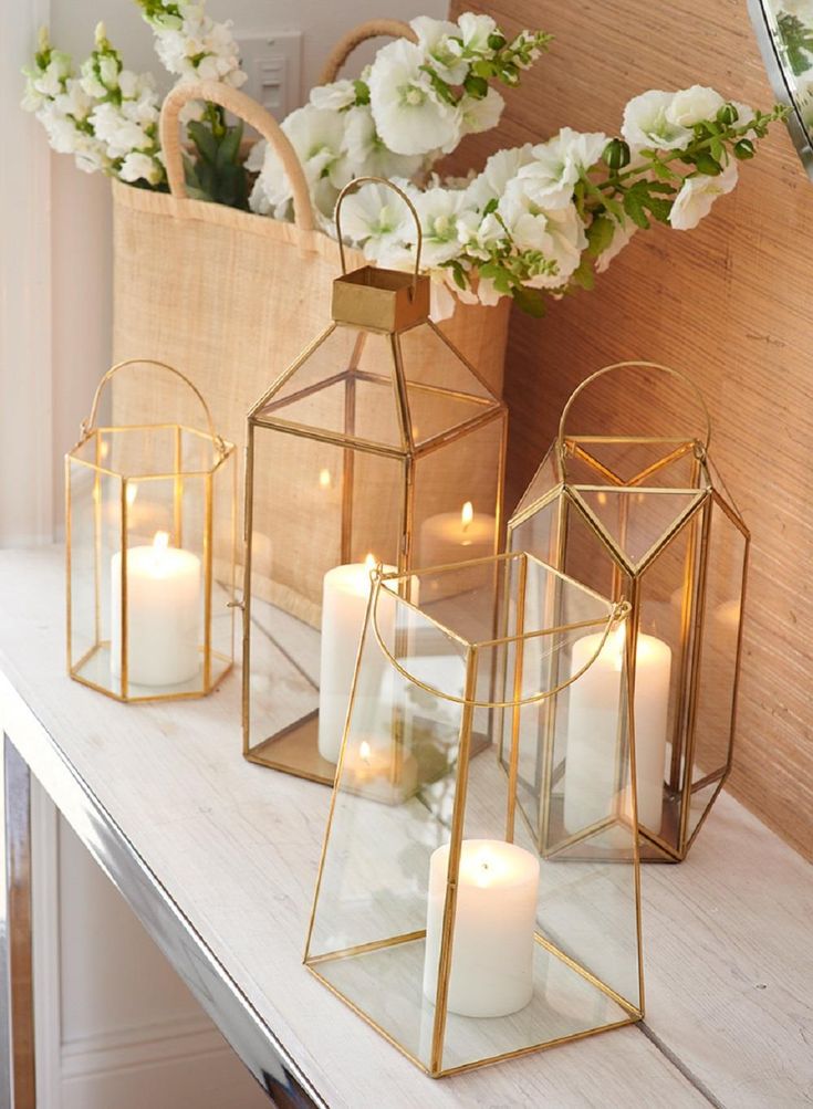 three gold lanterns with white flowers and candles on a table in front of a mirror