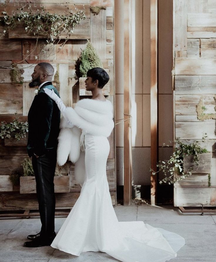 a man and woman standing next to each other in front of a wooden wall with greenery