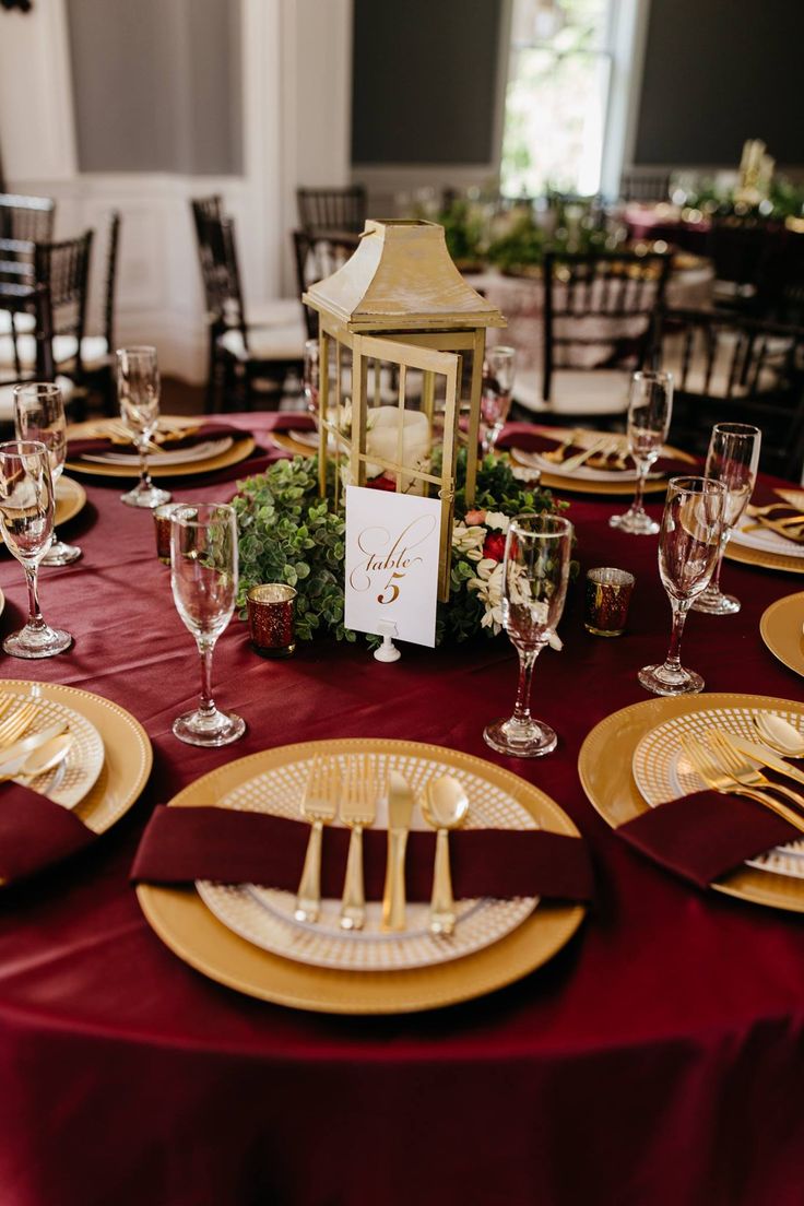 the table is set with gold and burgundy plates, place settings, and a lantern centerpiece