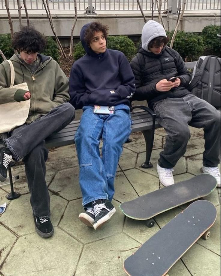 three boys sitting on a bench with their skateboards and looking at their phones while one boy is using his cell phone