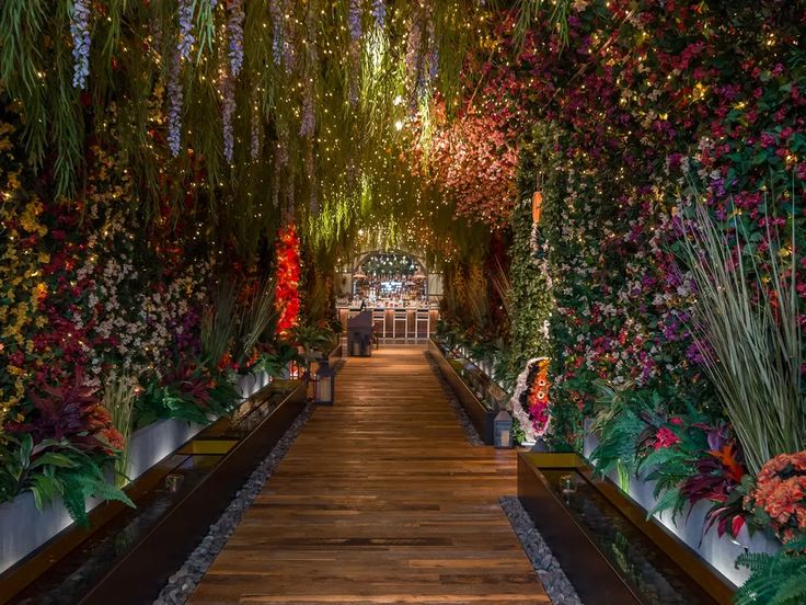 a walkway lined with lots of flowers and greenery next to a wall covered in lights