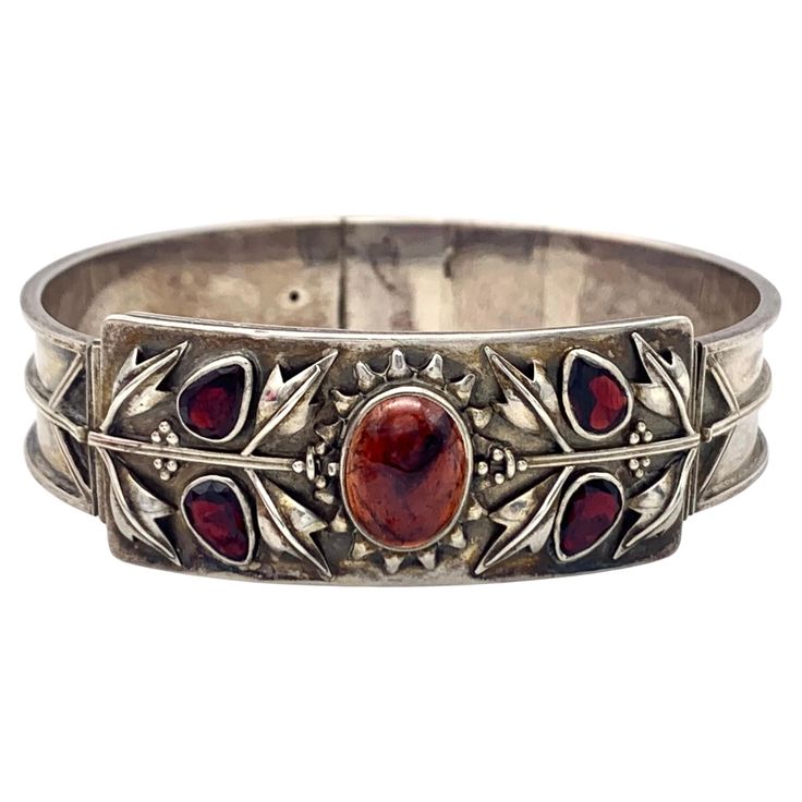 A large flower head with 16 flower petals and an oval cabochon embellishes the centre piece of this bracelet. The flower continues on either side into a flower stem with 6 leaves and small flower buds.. On each side we see two sets of silver leaves and a pair of garnet leaves with flat cut garnets. The knob of the clasp and the tongue of the bracelet have been gilt. The sides of the bracelet are decorated with a stylized tree in relief. The bracelet is photographed in the original patina, will h Stylized Tree, Cabochon Bracelet, Bracelet Flower, Silver Leaves, Flower Stem, Centre Piece, Flower Bud, Centre Pieces, Large Flowers