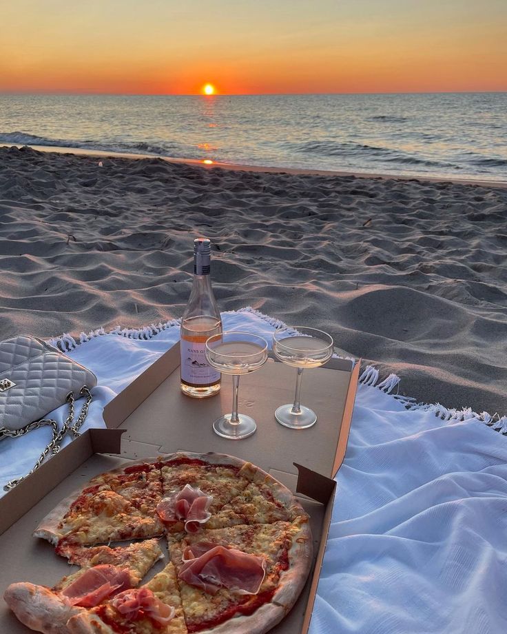 a pizza sitting on top of a beach next to a bottle of wine and two glasses